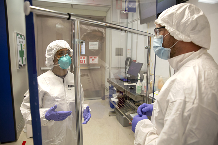 Two masked researchers discuss their work on opposite sides of a barrier made of a garment rack and a sheet of polycarbonate.