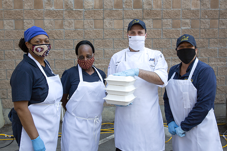 Cal Dining staff members in white aprons and face masks serve takeout lunches to students moving into the residence hall.