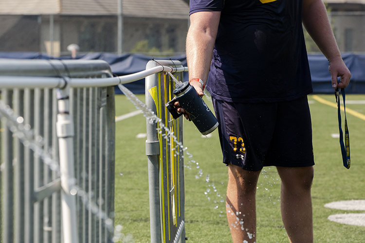 A football player fills his water bottle at a so-called water tree made of punctured PVC pipe attached to a hose. The water pours out every 6 feet, to keep players safe during the pandemic.