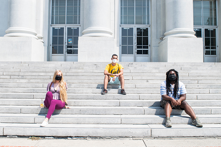 Three students who have just met sit in mask and 6 feet apart on the steps of Sproul Hall.
