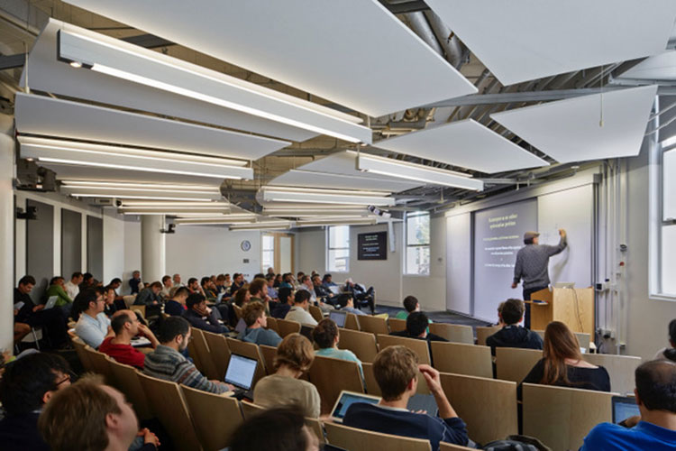 A photo of a large lecture hall in the Simons Institute. There is a person at the front of the hall talking and many people sitting in seats.