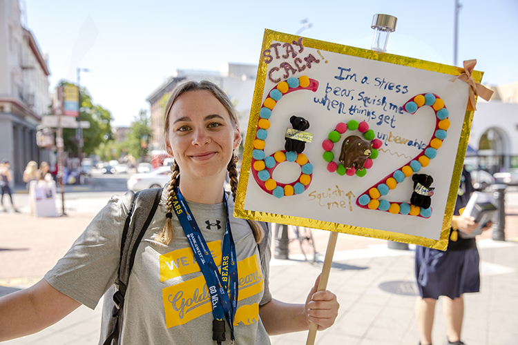 A student orientation leader for Golden Bear Orientation smiles and holds up a sign that says "Stay calm" and other slogans. She's on Sproul Plaza during GBO in 2019.