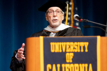 A photo of Geoffrey Nunberg at the 2019 I School Commencement. He is wearing academic regalia and standing behind a podium that says "University of California"