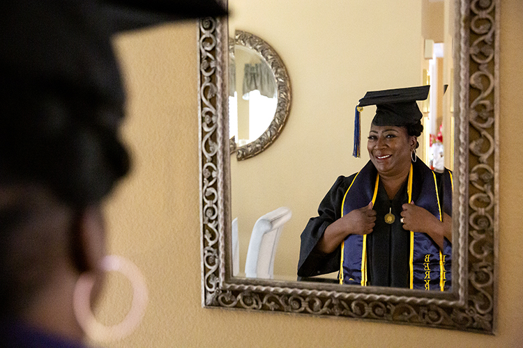 Jules Patrice Means stands in front of a mirror in her home wearing her regalia