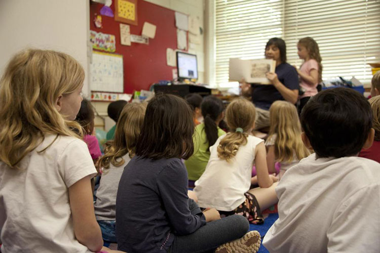 Young children sit on the floor, facing a women who is holding up a picture book for them to see