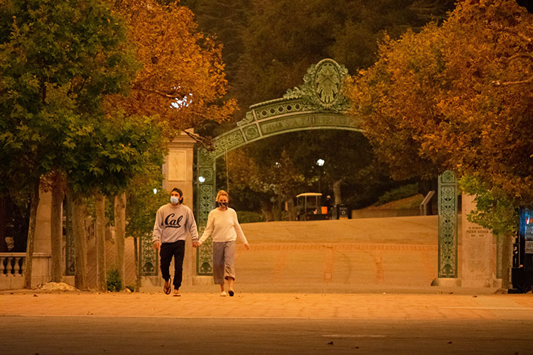 A couple holding hands and wearing masks walks across Sproul Plaza on a day when the skies were orange from Northern California wildfires.