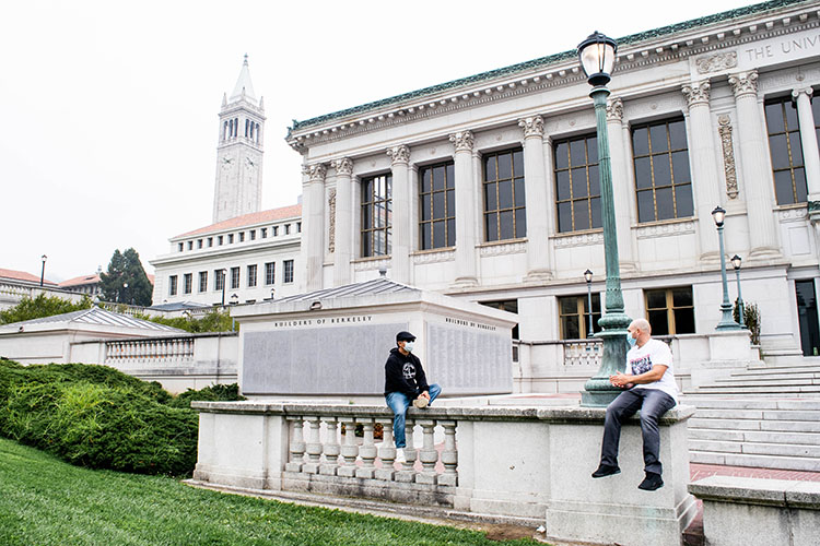 Henry Tsai, a Berkeley admissions officer, and Kevin McCarthy, a formerly incarcerated student, sit on a wall, many feet apart and wearing face masks, outside of Doe LIbrary.