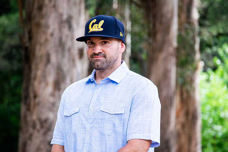 Kevin McCarthy, a formerly incarcerated student, wears a Cal cap and stands in front of a grove of eucalyptus trees on campus.