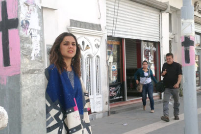 Laila stands on a street next to two poles with crosses painted on them