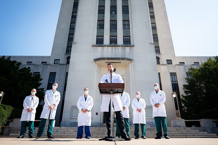 Seven doctors in white lab coats talking at a pdium in front of a hospital building