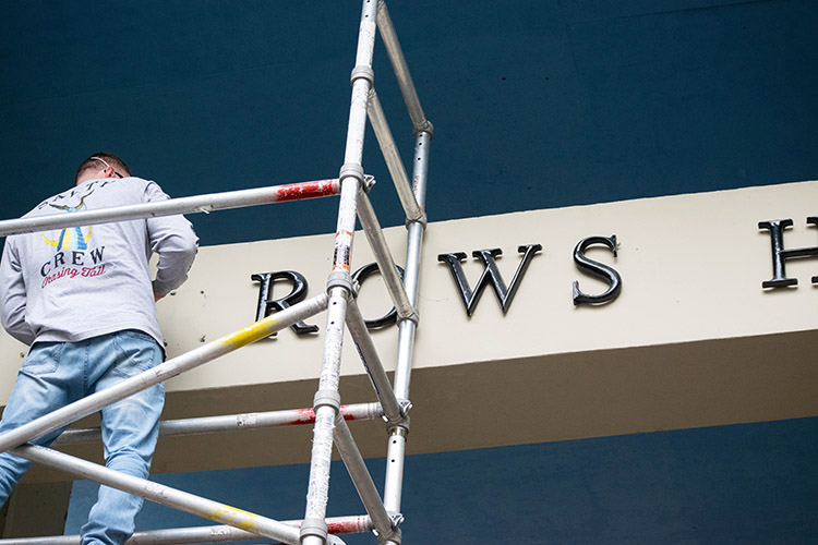 A worker stands on scaffolding to remove metal letters from the exterior of Barrows Hall.