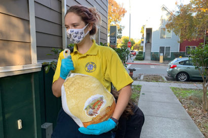 A person wearing a yellow shirt, a face mask and gloves carries a large frozen turkey.