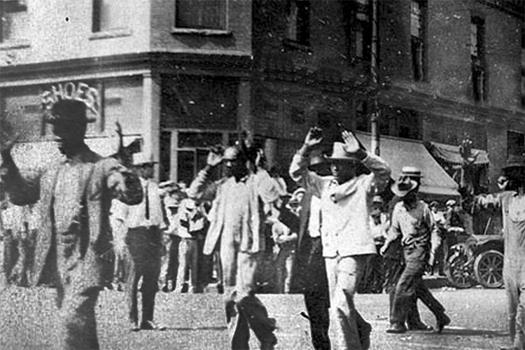 Black men, some with their hands up, are forced by the National Guard to walk through the streets of Tulsa during the Tulsa Race Massacre of 1921.