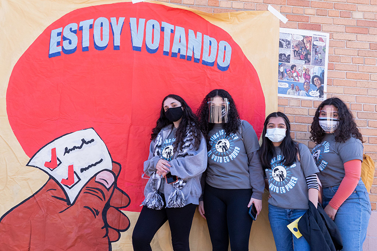 four people pose by a mural that reads "Estoy votando"