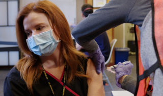 A nurse with red hair gets the COVID-19 vaccine while wearing a mask