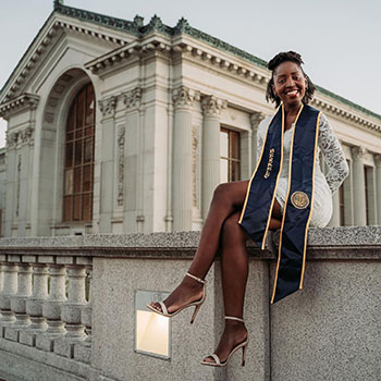 Gloria Tumushabe poses in a graduation stole that she wore when she received her undergraduate degree from Berkeley. She is sitting on a ledge near University Library.