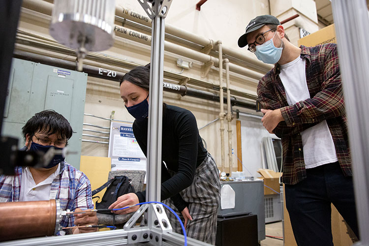 A photo shows three researchers gathered around a gold cylinder inside an open lab space