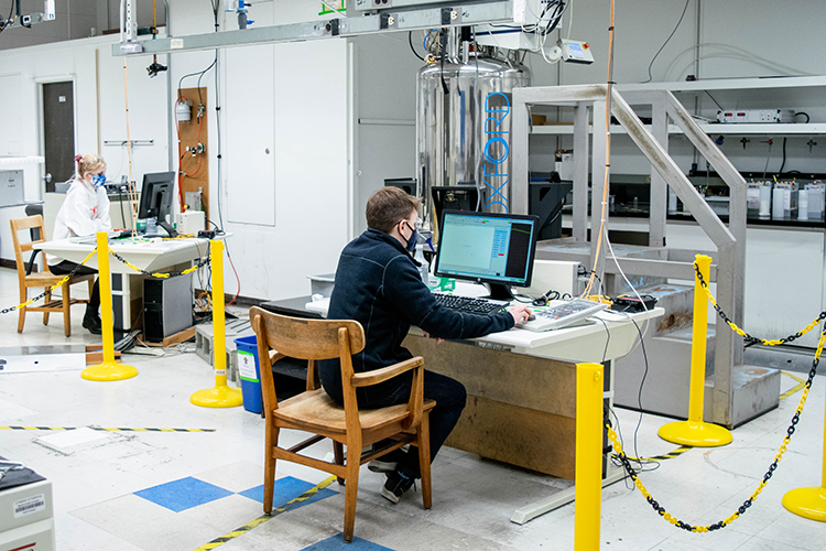 Two graduate students in masks sit at computers in the Nuclear Magnetic Resonance Facility in Latimer Hall. They are separate by stanchions and heavy plastic chains, part of precautions taken during the pandemic.