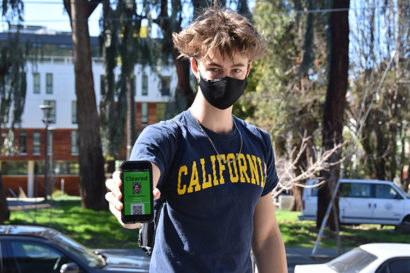 Student Jonas Willett, wearing a face mask and a California T-shirt, shows his cell phone screen to a health ambassador outside Crossroads dining commons. The screen is green, indicating that he is current with his required COVID-19 testing on campus.