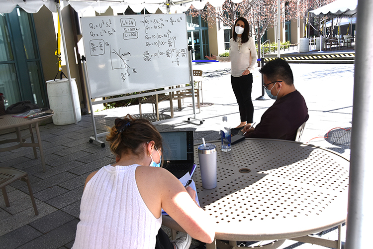 A Berkeley Haas associate professor leads an outdoor session for a handful of undergraduates in her international trade course under a tent on the Berkeley Haas courtyard. She is wearing a mask and standing at a whiteboard.