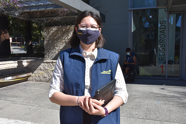 Gabriele Mackie stands in front of Crossroads dining hall wearing a student ambassador vest and a black face mask. She is supervising the health ambassadors who check students' e-badges to show they've been cleared to enter UC Berkeley buildings like Crossroads.