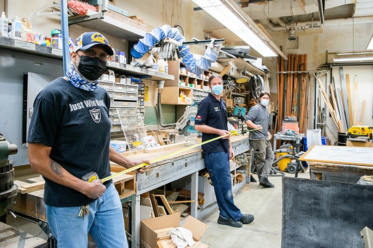 The team of three men who run the Sheet Metal and Wood Shop on campus are all essential workers who have been here daily during the pandemic. They stand, wearing masks, in a row, holding a measuring tape to show 6 feet between them.