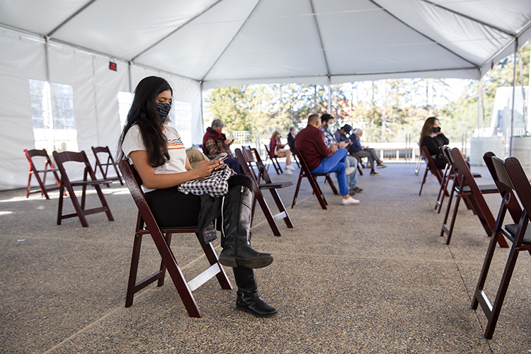 After people get vaccines in Pauley Ballroom, they go outside onto the third floor deck and into an open air tent, where they are monitored for vaccine reactions by members of the Berkeley Medical Reserve Corps. In this photo, people sit in folding chairs, socially distanced from each other, after their vaccinations.