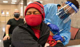 A woman wearing a mask flinches as a health care working wearing a face shield administers a shot into her left arm
