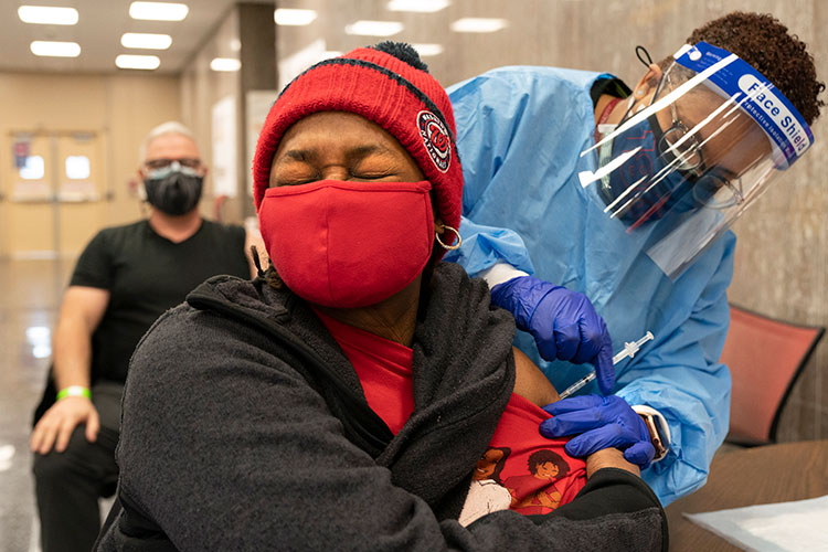 A woman wearing a mask flinches as a health care working wearing a face shield administers a shot into her left arm