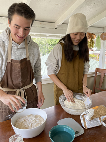 Fellow bread bakers Brendan Huang and Carolyn Hong wwork together in a kitchen where they are making sourdough. They're wearing aprons.