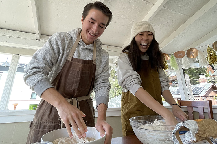 Students Brendan Huang and Carolyn Hong bake sourdough bread together. They're wearing aprons and laughing.