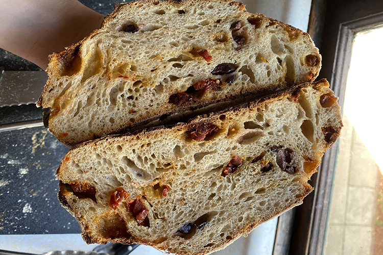 A loaf of sourdough bread, cut in half, reveals sun-dried tomatoes and olives.