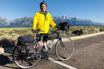 Harley Shaiken poses with his bike with the Teton Range in the background. He's wearing a bright yellow parka andblack biking shorts.