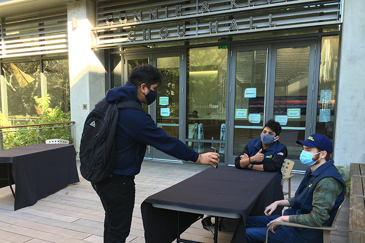 Two student health ambassadors sitting at a table outside Chou Hall check a student's cell phone for proof that he recently was tested for COVID-19.