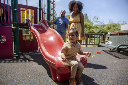 a young boy on a slide on a playground with his two parents watching