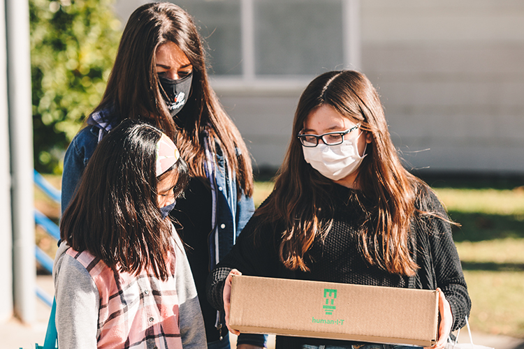 Three girls in masks during the pandemic walk away from a technology giveaway with a box containing a free laptop made possible by Connect-In-Place, a free remote camp that also raised money for students in need of laptops and other technology.