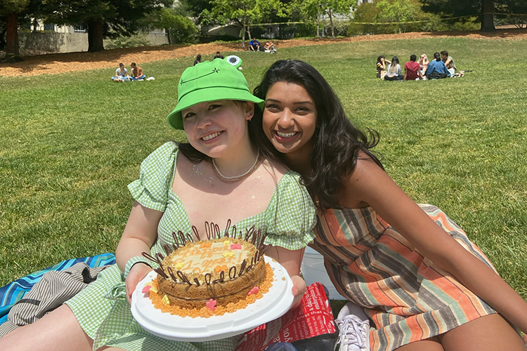 Danielle Egan and Saumya Goyal, co-founders of the Connect-In-Place online camp for youth, sit together on Memorial Glade smiling with a cake Egan made to celebrate the camp's first anniversary.