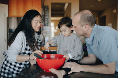 A woman, a man, and a small boy gather around a mixing bowl in a kitchen.
