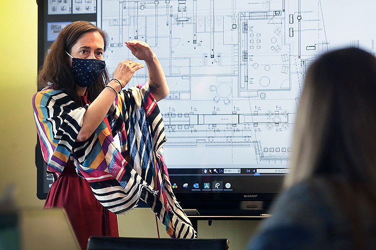 woman standing before a projection of an architectural drawing, gestures to a seated colleague