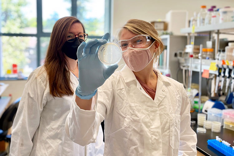 Two scientists stand in a lab, wearing lab coats and goggles. One is holding up a Petri plate.