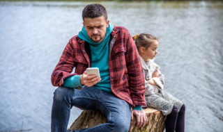 Adult frowning at smartphone while child sits with arms folded.