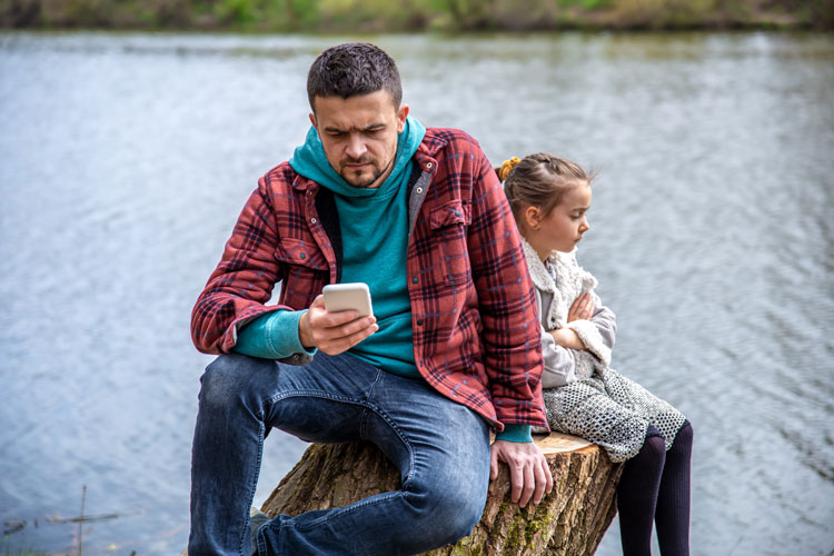 Adult frowning at smartphone while child sits with arms folded.