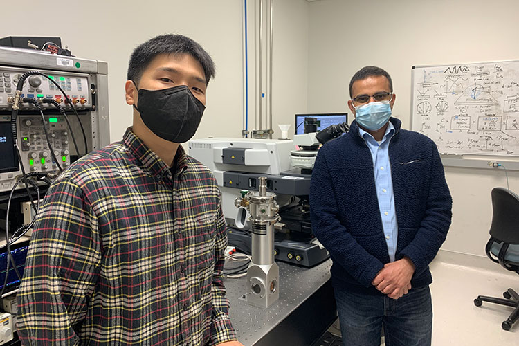 A photo shows two people standing in an optical laboratory. Both are wearing face masks.