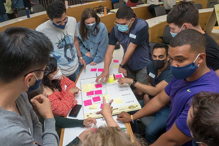 First-year Berkeley Haas MBA students in masks participate in a design/think session during orientation week. They are gathered around a table filled with post-it notes and are talking and pointing to them.