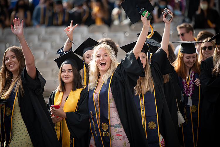 A group of young women in regalia smile and wave at the Class of 2020 In-Person Commencement at the Greek Theatre.