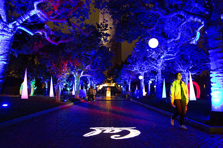 Students walk through a light display shining on the trees and pavement next to the Campanile during Golden Bear Orientation's Paint the Night event. The word Cal in white lights appears on the sidewalk and the trees are shades of bright blue.