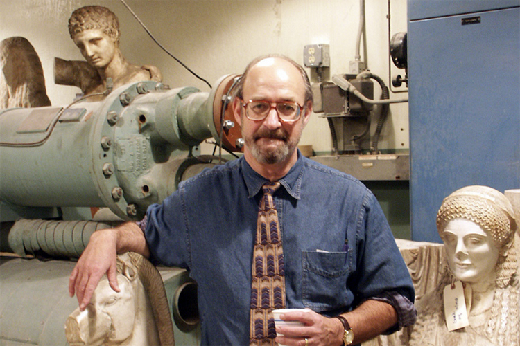 Stephen Miller, a professor emeritus of classics who died Aug. 11, 2021, stand in front of casts of ancient statuary holding a styrofoam coffee cup and looking at the camera.