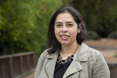 New assistant professor Danielle Zoe Rivera smiles for the camera in close-up shot taken in a wooded area of campus.