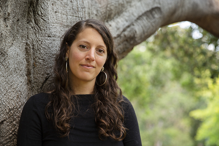 New assistant professor Meg Mills-Novoa leans against a tree and looks at the camera.