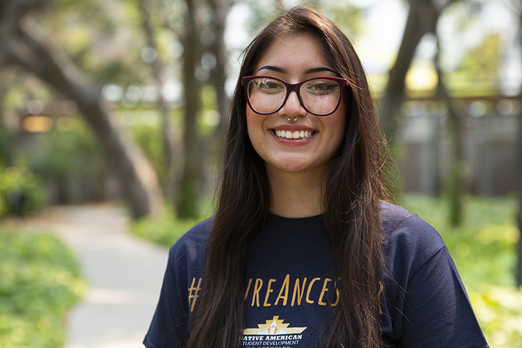 First-year student Sariel Sandoval, who is Native American, poses for a portrait on campus wearing a blue and gold Berkeley T-shirt and large, stylish eyeglasses.